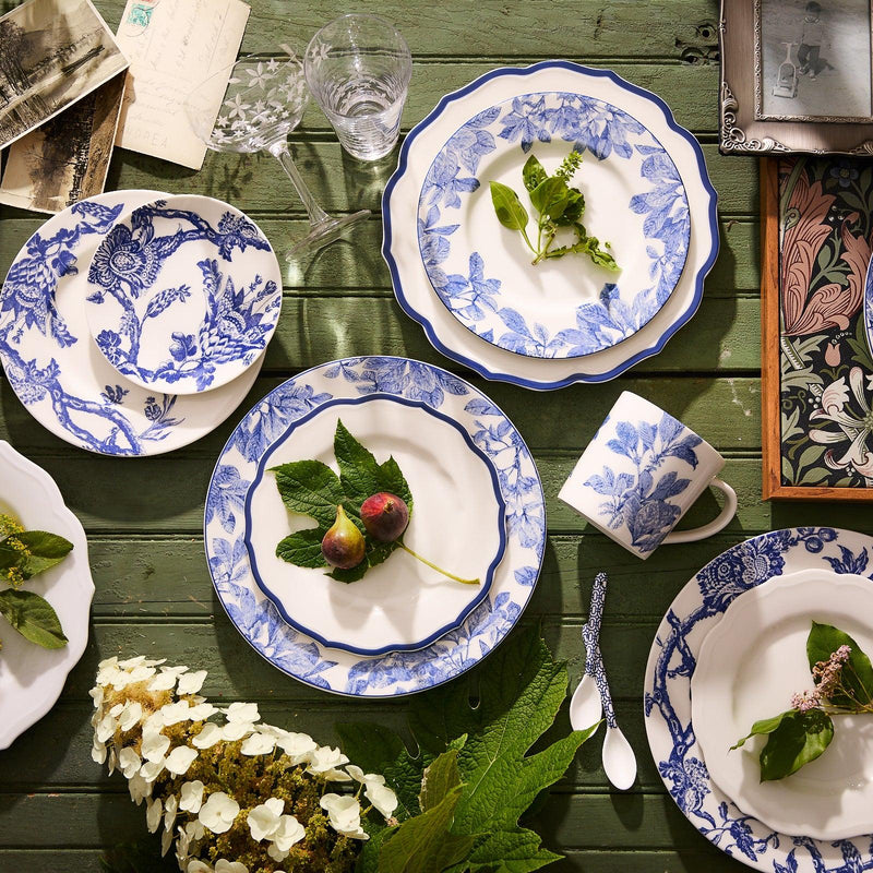 A green wooden table set with blue and white floral-patterned premium porcelain plates, an Arbor Rimmed Salad Plate by Caskata Artisanal Home, a cup, cutlery, a wine glass, leaves, and figs. Nearby are a bouquet, old photographs, and a patterned notebook. All items are conveniently dishwasher safe.