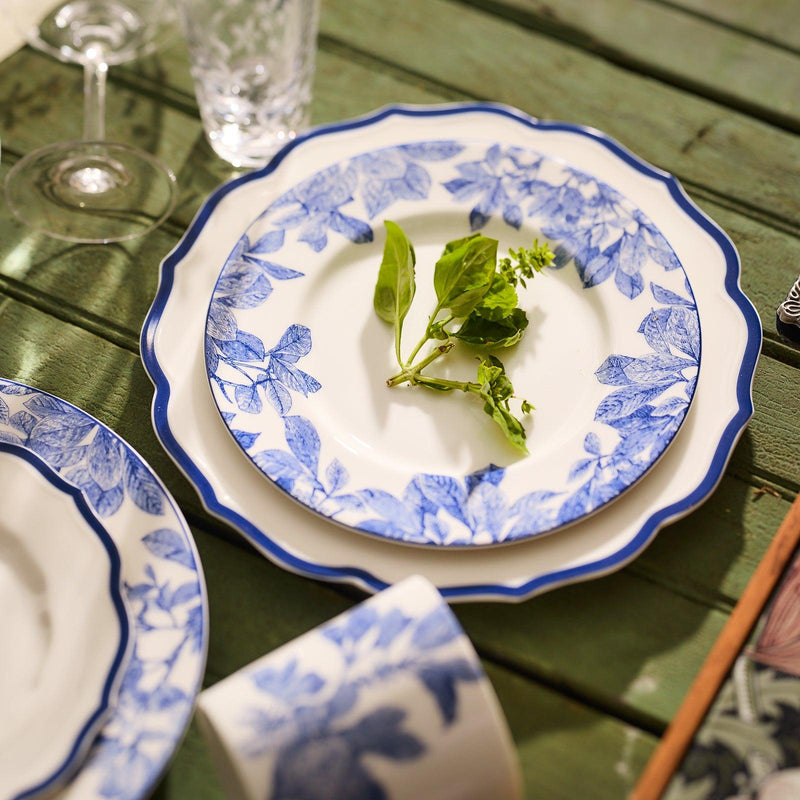 An Arbor Rimmed Salad Plate from Caskata Artisanal Home, featuring white porcelain adorned with blue floral designs, holds a few green sprigs. It rests on a wooden table alongside additional plates and a glass nearby, enhancing the rustic charm of the setting with its premium porcelain craftsmanship.