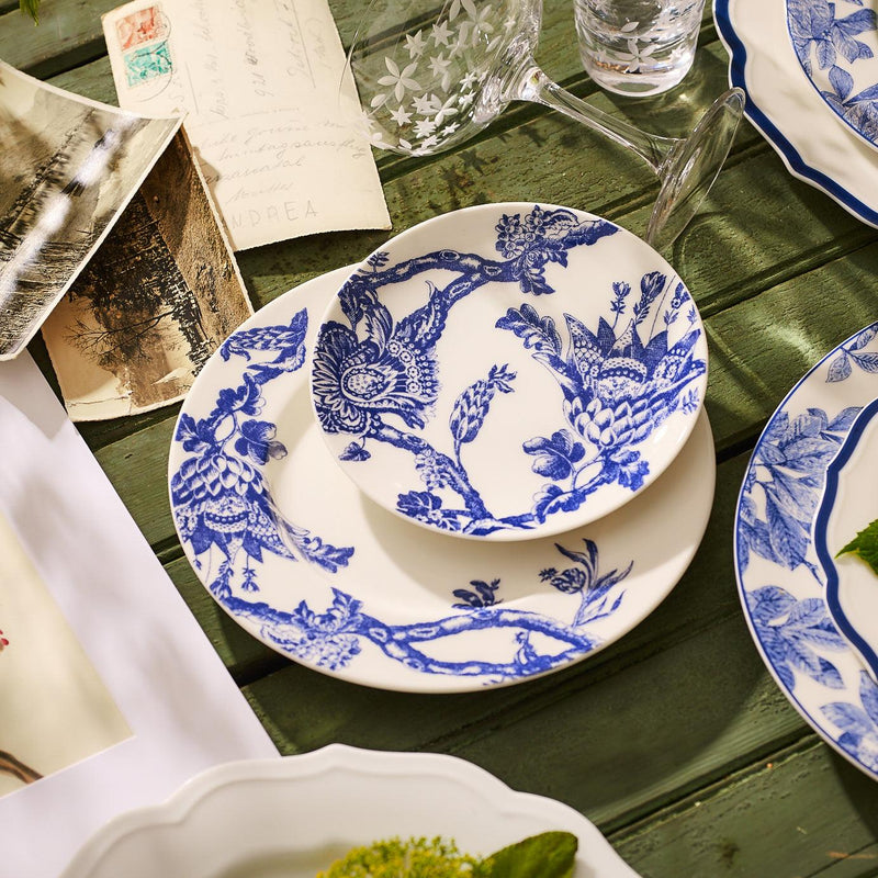 A set of Arcadia Rimmed Salad Plates by Caskata Artisanal Home, crafted from premium porcelain and featuring intricate floral and bird designs in blue-and-white, sits on a wooden table surrounded by letters and a glass of water. This elegant collection brings an heirloom feel to any setting.