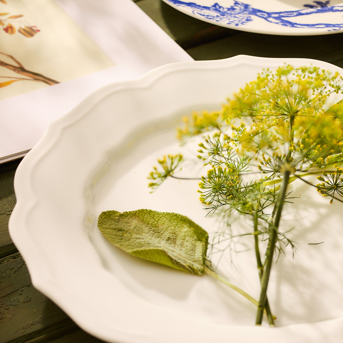 The Stella Scalloped White Dinner Plate by Caskata, adorned with a green leaf and yellow flowers, exudes a vintage feel. It sits elegantly beside an open book of botanical illustrations, seamlessly harmonizing with the neutral palette of timeless dinnerware patterns.