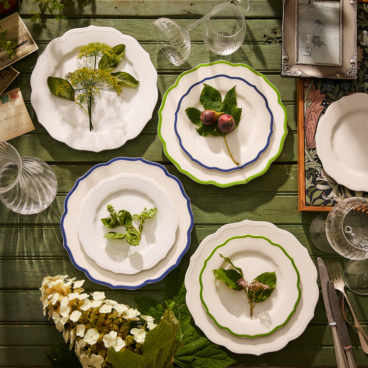 On a wooden table, Caskata&#39;s Stella Scalloped Verde Dinner Plates, adorned with intricate scalloped details, serve as the backdrop for an arrangement of leaves and flowers. The scene is complemented by heirloom-quality porcelain glassware, a knife, and scattered photos, creating a charmingly nostalgic atmosphere.