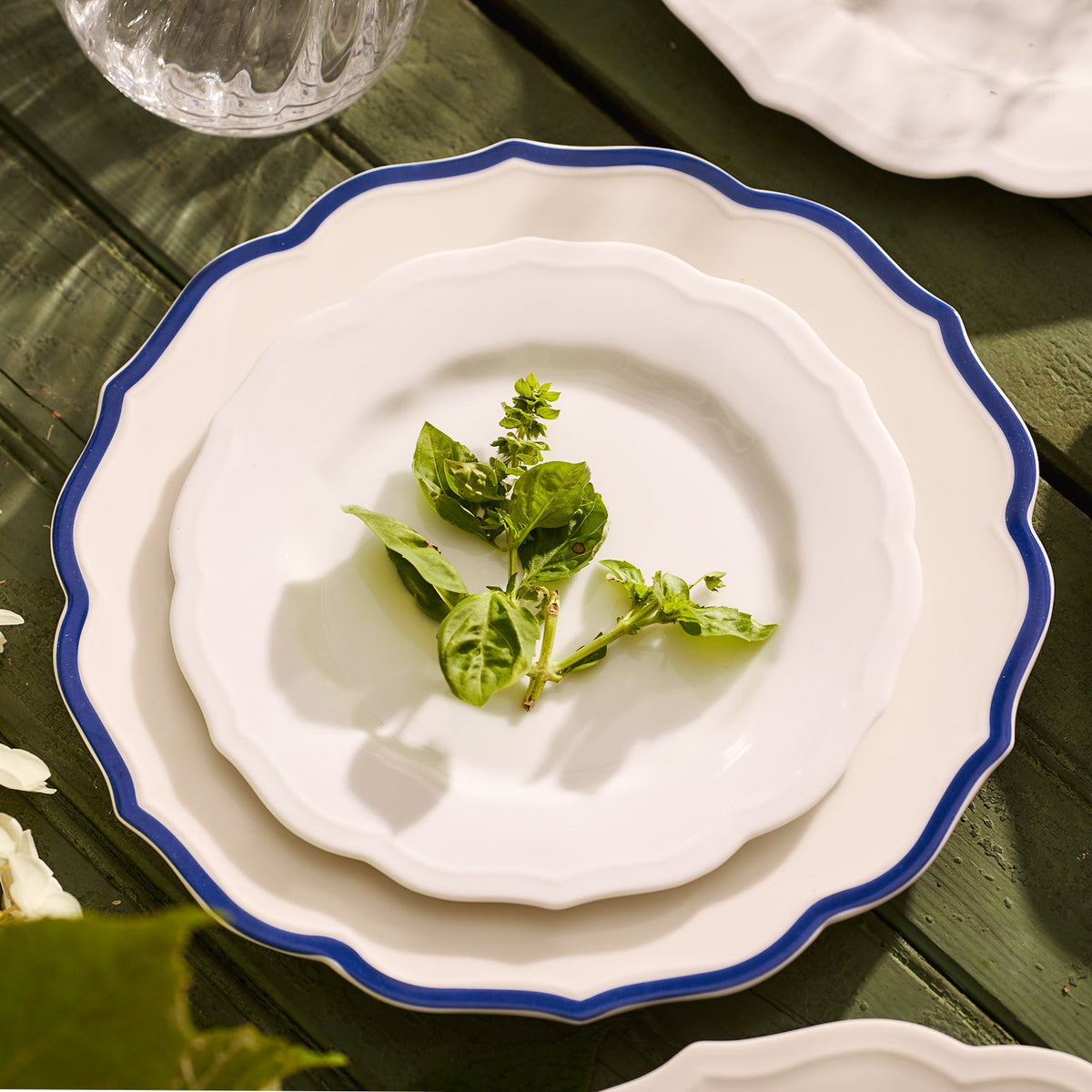 A stack of three Stella Scalloped Blue Dinner Plates by Caskata, resting on a green wooden table with a vintage vibe, are adorned with fresh basil sprigs.
