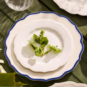 A stack of three Stella Scalloped Blue Dinner Plates by Caskata, resting on a green wooden table with a vintage vibe, are adorned with fresh basil sprigs.