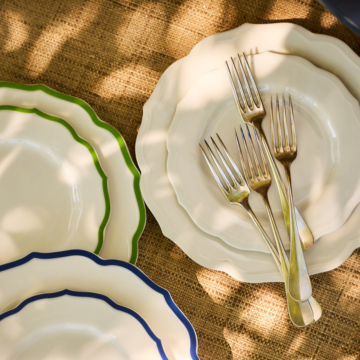A set of Caskata&#39;s Stella Scalloped White Dinner Plates with green and blue rims, accompanied by a stack of forks on a burlap surface, creates a vintage ambiance as dappled sunlight casts shadows.