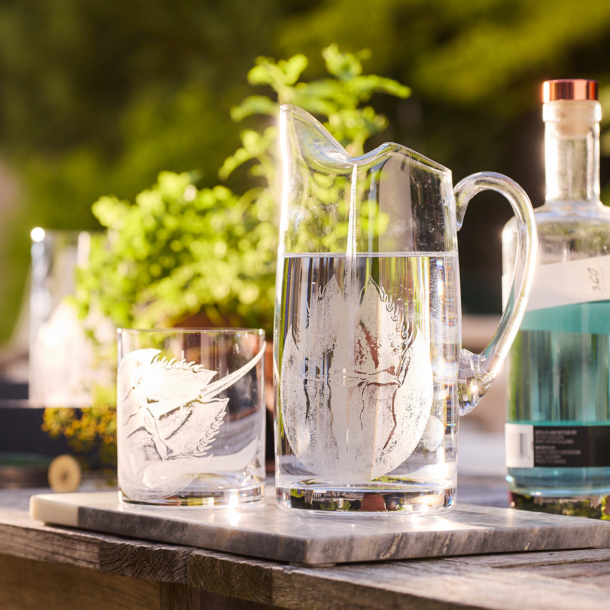 A glass pitcher and tumbler, named the Horseshoe Crab Small Pitcher by Caskata, crafted from lead-free crystal using a delicate sand-etching technique to display intricate leaf designs, sit on an outdoor table next to a bottle of blue liquid, with lush greenery in the background.