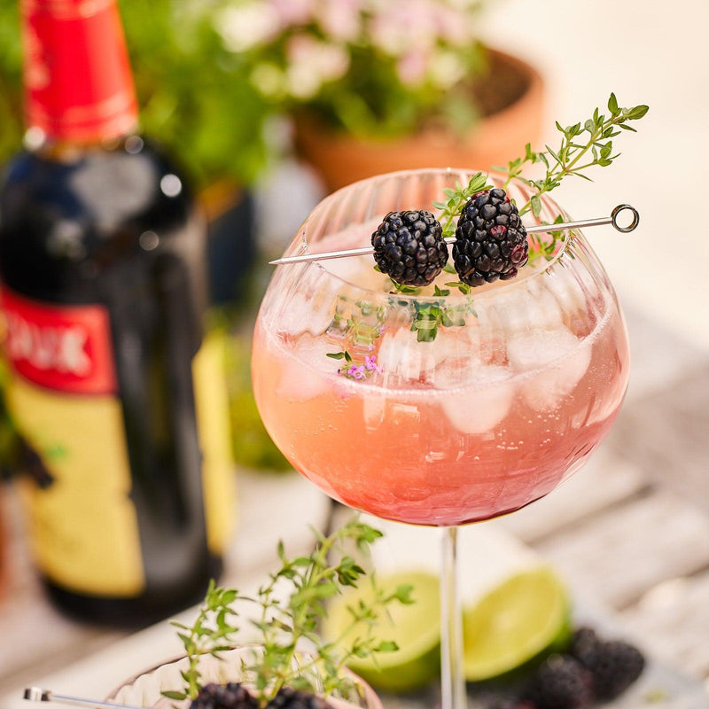 A pink cocktail garnished with blackberries and thyme in a mouth-blown Quinn Clear Red Wine Glass from Caskata Artisanal Home, with a bottle and lime wedges in the background on a wooden table.