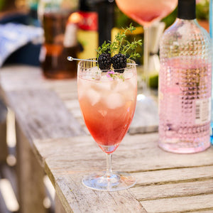 A Quinn Clear Everyday Glass by Caskata Artisanal Home, mouth-blown and crafted from lead-free crystal, is filled with a pinkish drink, ice cubes, and garnished with blackberries and a sprig of greenery. The exquisite Czech Republic glassworks piece rests on a wooden table amidst other bottles and glasses in the background.