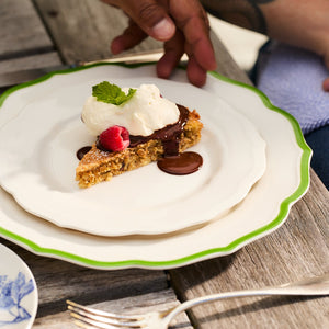 A slice of dessert on a Stella Scalloped Verde Dinner Plate by Caskata, topped with cream, chocolate sauce, a raspberry, and mint. A hand is visible, and a fork rests nearby on the wooden table.