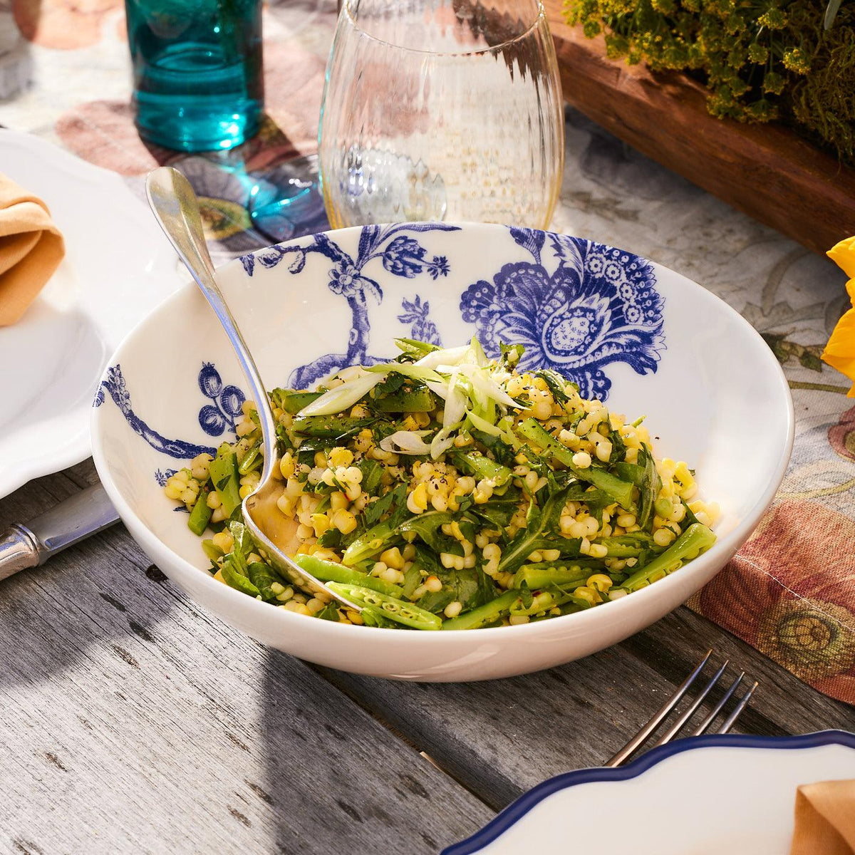 A bowl of couscous salad with vegetables is placed on an outdoor table setting, showcased within the premium Arcadia Entrée Bowl by Caskata Artisanal Home, accompanied by a fork, knife, and glasses.