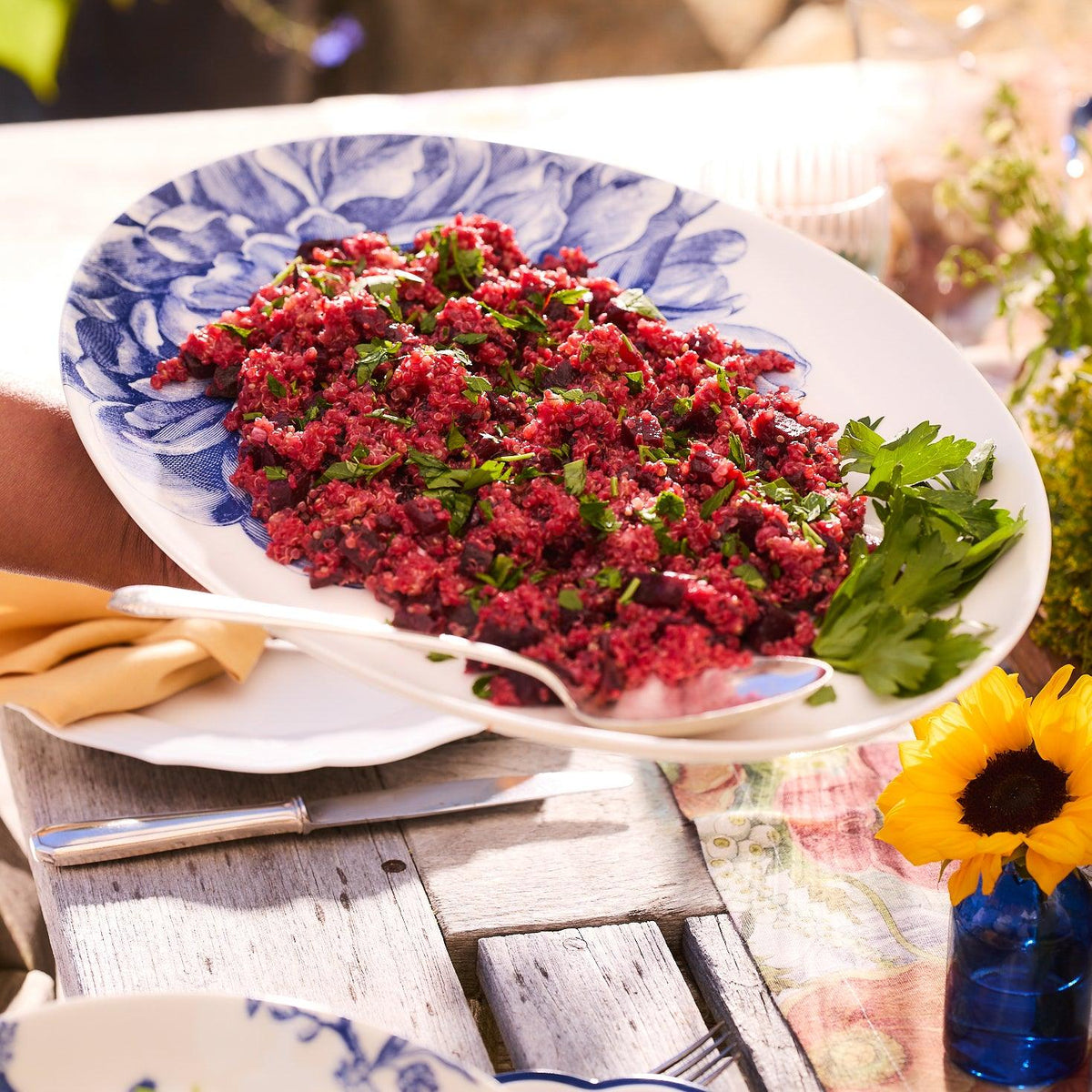 A plate of quinoa and beet salad garnished with fresh herbs, served on a Peony Medium Coupe Oval Platter from Caskata Artisanal Home, with a napkin and sunflower decoration on a rustic wooden table.