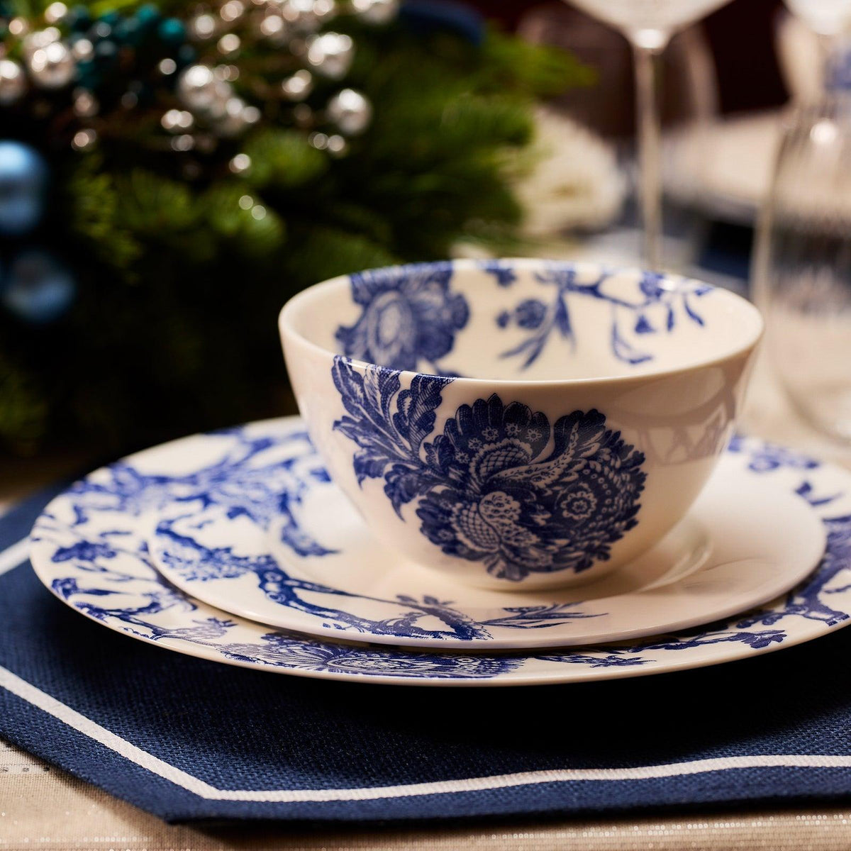 A white Caskata Arcadia Cereal Bowl and plates adorned with blue floral patterns are stacked on a table, placed on a blue and white napkin. A green centerpiece with blue and silver ornaments is blurred in the background, completing the elegant Arcadia porcelain dinnerware setting.