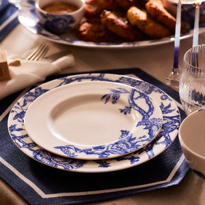 An array of Arcadia Rimmed Salad Plates by Caskata Artisanal Home, featuring a beautiful blue and white pattern and crafted from premium porcelain that feels like heirlooms, nestled together on a blue placemat alongside a glass, cutlery, and a side dish on the dining table.