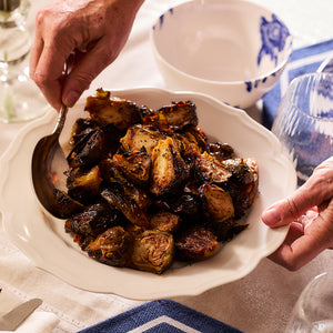 A person serves roasted Brussels sprouts on a Stella Scalloped White Dinner Plate by Caskata, with its subtle patterns adding a touch of elegance to the table.