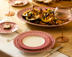 A table set with Caskata's Casablanca Crimson Table for 12—red and white plates, cutlery, a pink wine glass, and roasted vegetables on a beige and white tablecloth.