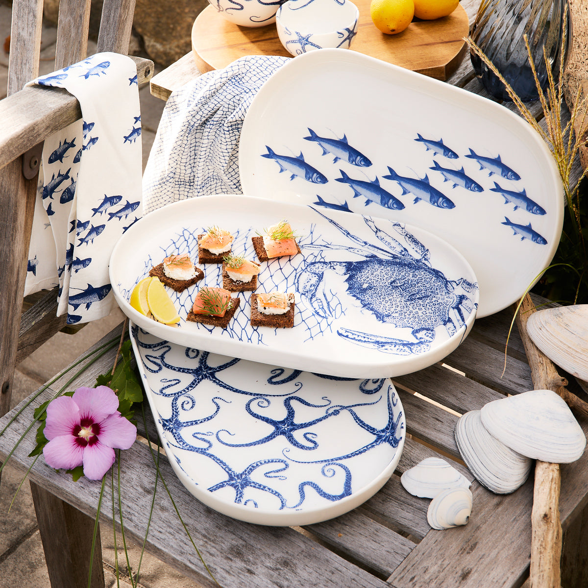 Caskata&#39;s Starfish Large Coupe Oval Platter with blue marine designs of fish and octopuses rests elegantly on a wooden chair. Some platters display appetizers alongside a lemon wedge and a purple flower. Nearby, seashells, starfish, and a towel create an ideal setting for ocean-themed entertaining.