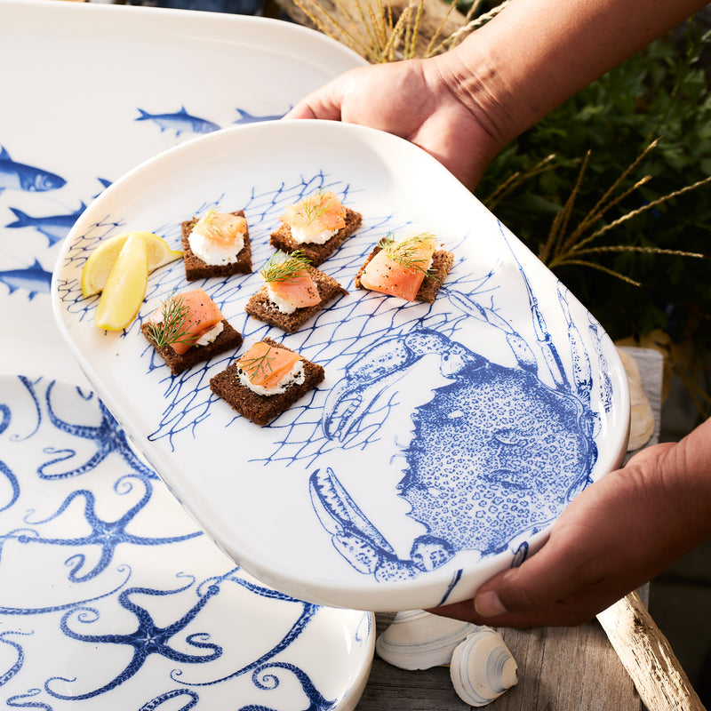 Hands offering an entertaining platter adorned with salmon-topped crackers, garnished with lemon wedges. The Crab Large Coupe Oval Platter by Caskata is crafted from high-fired porcelain and features a blue nautical crabs pattern alongside a fish design.