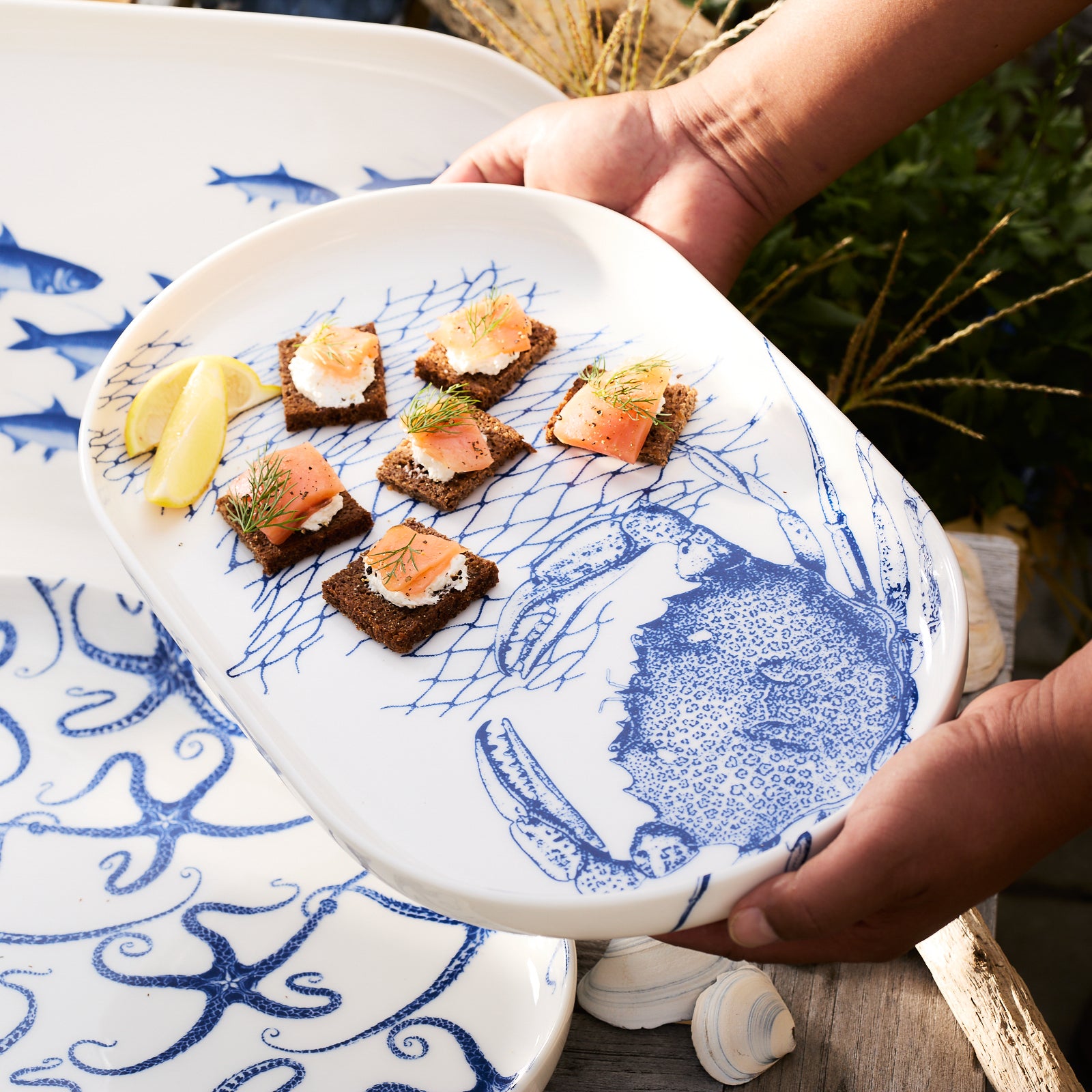 The Crab Large Coupe Oval Platter by Caskata features a charming blue illustration of a crab and fishing net design. Crafted from high-fired porcelain, this piece beautifully captures the crabs pattern, making it perfect for serving with style.