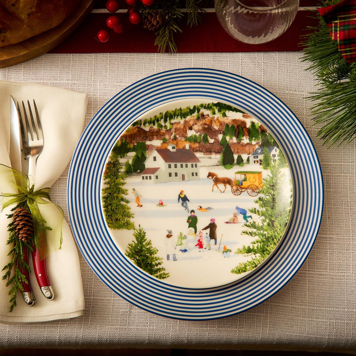 A Newport Stripe Rimmed Dinner Plate by Caskata Artisanal Home, featuring a winter village scene with people ice skating and a horse-drawn carriage, is elegantly set on the table. Beside it, a fork and knife wrapped in a napkin complete the festive setting.
