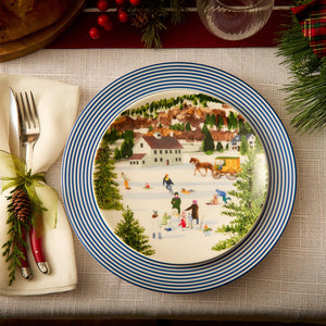 A Newport Stripe Rimmed Dinner Plate by Caskata Artisanal Home, featuring a winter village scene with people ice skating and a horse-drawn carriage, is elegantly set on the table. Beside it, a fork and knife wrapped in a napkin complete the festive setting.