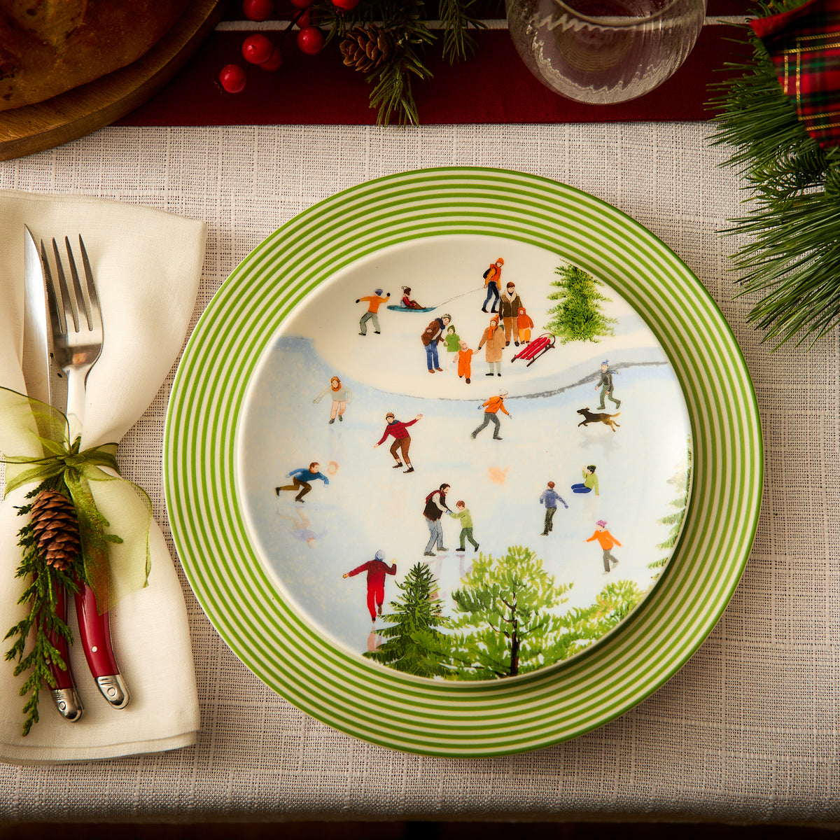 A festive holiday table setting features a high-fired porcelain dinner plate with a winter ice skating scene, placed on a Caskata Artisanal Home Newport Stripe Verde Rimmed Dinner Plate, accompanied by a cloth napkin with silverware and a pinecone decoration.
