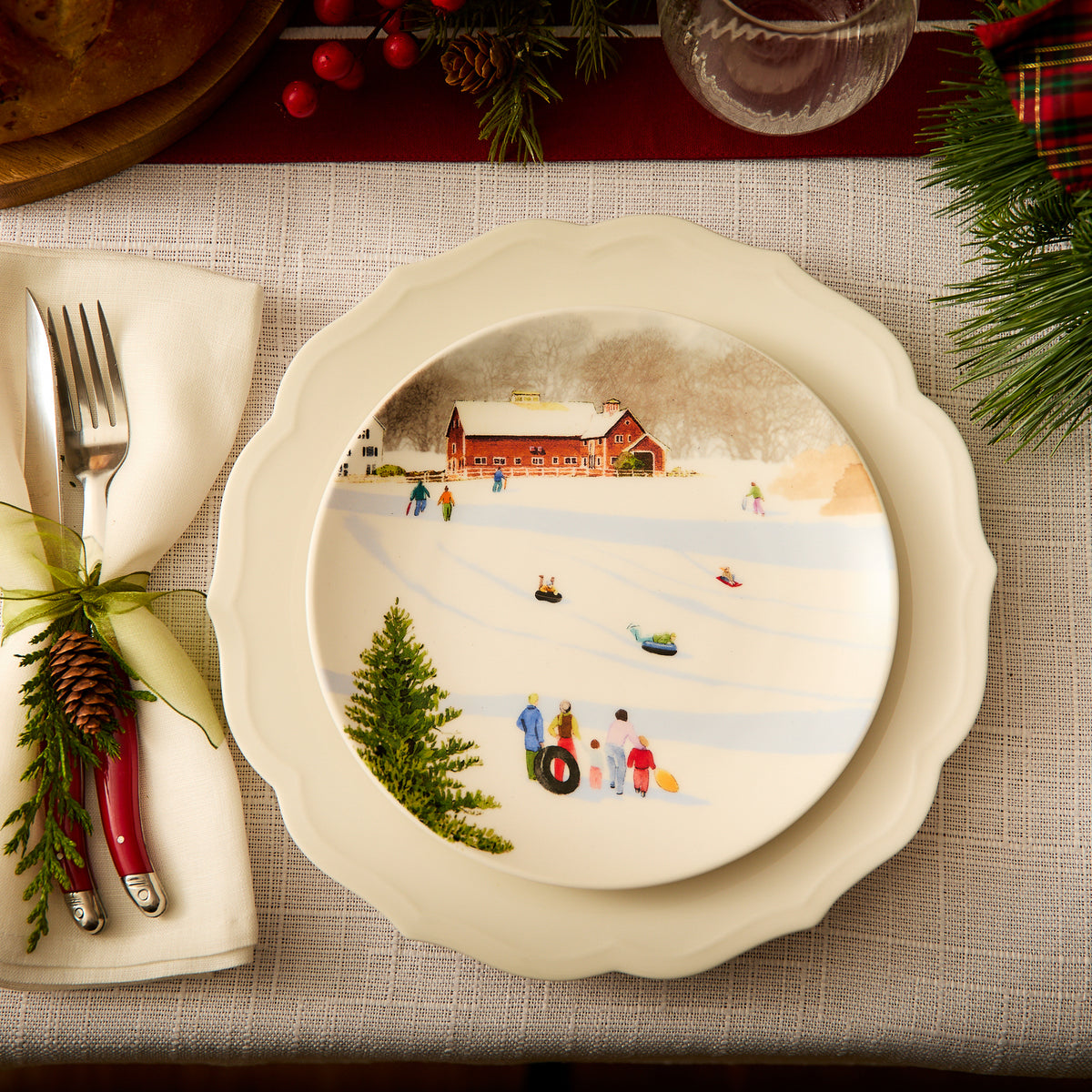 Create a festive dinner table setting with a vintage charm by featuring the Stella Scalloped White Dinner Plate from Caskata, which beautifully captures a winter scene of people sledding near a red barn. The silverware is elegantly wrapped in a napkin adorned with a pine cone and green ribbon, perfectly complementing the classic patterns of this elegant dinnerware.