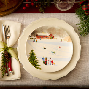 Create a festive dinner table setting with a vintage charm by featuring the Stella Scalloped White Dinner Plate from Caskata, which beautifully captures a winter scene of people sledding near a red barn. The silverware is elegantly wrapped in a napkin adorned with a pine cone and green ribbon, perfectly complementing the classic patterns of this elegant dinnerware.