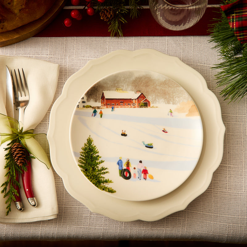 A Caskata X Felix Doolittle Winter Wonderland Plate, featuring an illustration of people sledding in the snow reminiscent of Felix Doolittle’s watercolor landscapes, is placed on a white tablecloth with a napkin, cutlery, and Christmas greenery decor nearby.