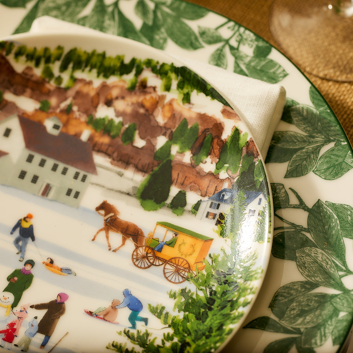 A decorative porcelain salad plate from the Caskata X Felix Doolittle Winter Wonderland Plates, Set of 4, features a winter village scene with people ice skating and a horse-drawn carriage, bordered by Felix Doolittle&#39;s signature leafy green patterns and set on a table with a napkin and another plate underneath.