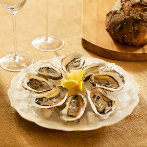 A vintage scene is evoked by a Stella Scalloped White Dinner Plate from Caskata, filled with oysters on ice and garnished with lemon wedges. It sits beside two empty wine glasses and a loaf of bread on a wooden board, creating a charming atmosphere with its neutral palate.