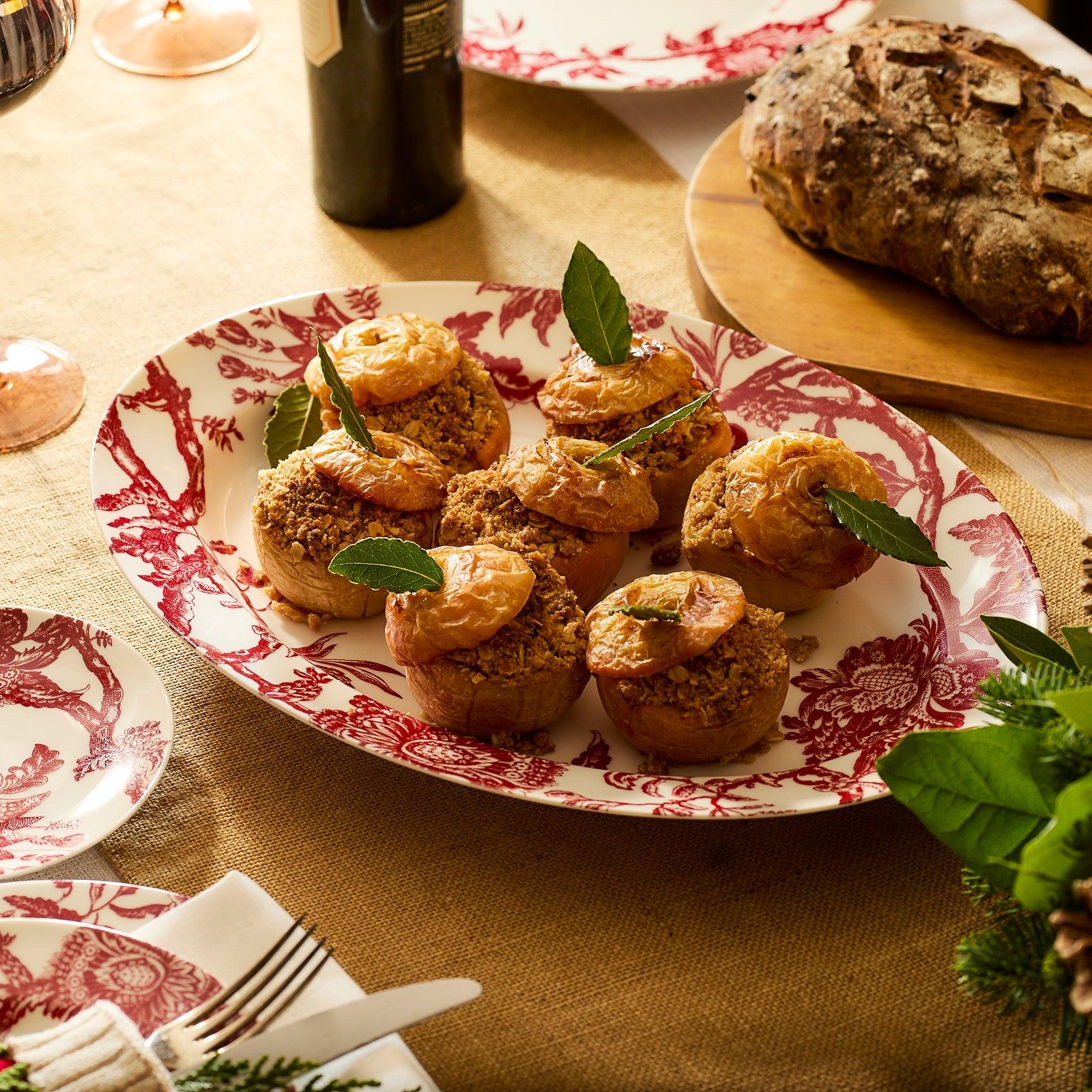 Caskata’s “Happy Hosting” Bundle features oval and round Arcadia Crimson plates with red floral designs, styled with a yellow ribbon, scissors, pen, gift tags, and an oversized platter for a festive presentation.