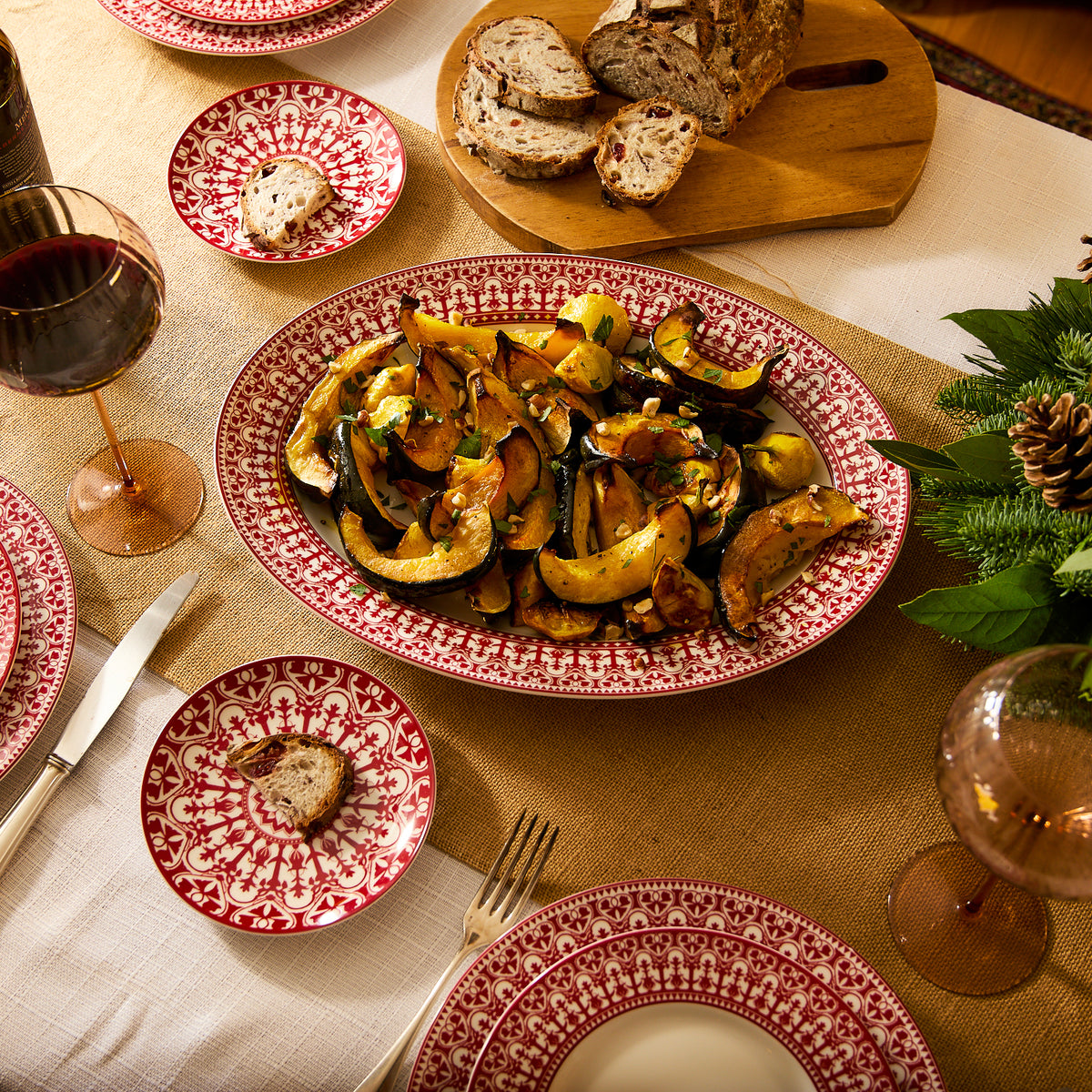 A dining table set with Casablanca dinnerware, red-patterned plates, wine glasses, cutlery, and a Casablanca Crimson Oval Rimmed Platter by Caskata Artisanal Home filled with roasted vegetables. Sliced bread rests on a cutting board while a small plate holds a slice of bread and a pat of butter.