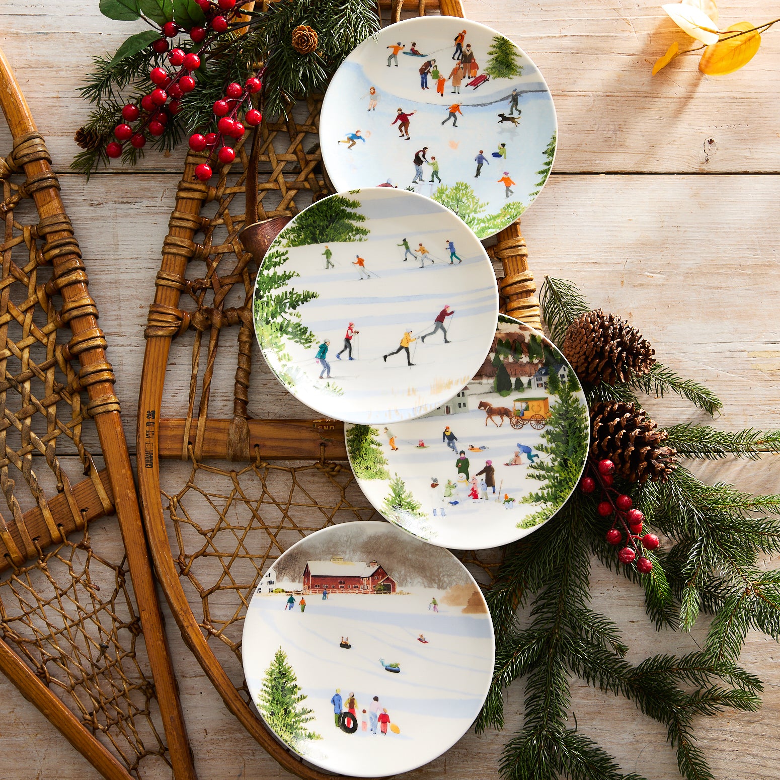 A set of four Caskata X Felix Doolittle Winter Wonderland Plates by Caskata, featuring watercolor landscapes of New England winter scenes with people sledding, skating, and a horse-drawn sleigh in snowy settings with trees, buildings, and mountains in the background.