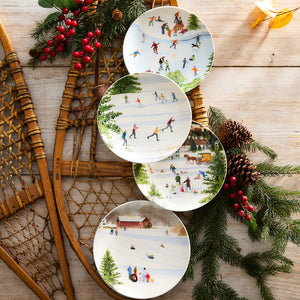 The Caskata X Felix Doolittle Winter Wonderland Plates, a set of four featuring watercolor winter scenes of people ice skating, skiing, and enjoying snowy landscapes by Felix Doolittle, are arranged on a wooden surface alongside vintage snowshoes and pine decorations.