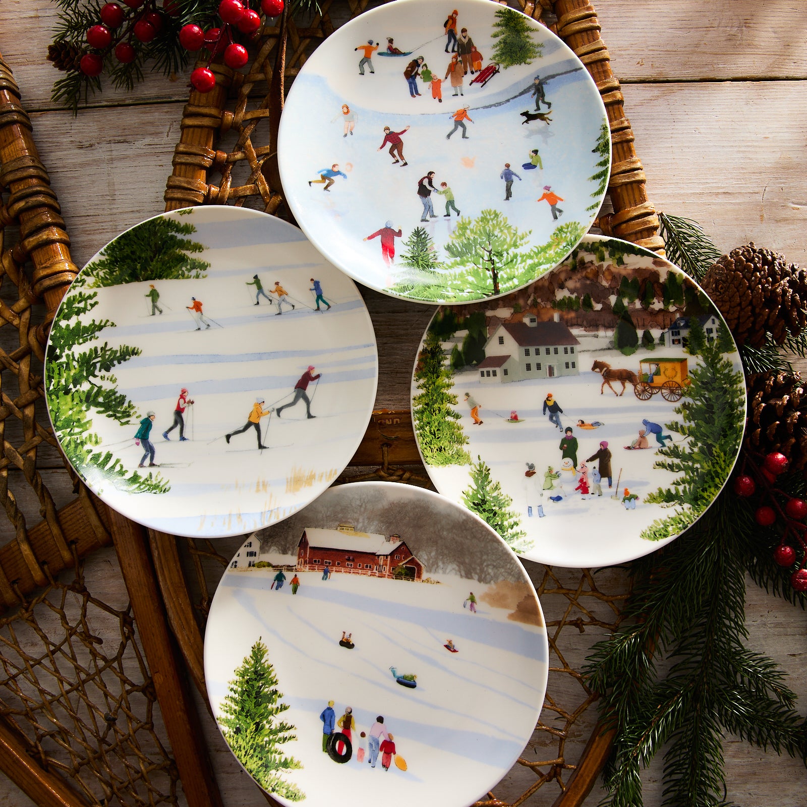 A set of four Caskata X Felix Doolittle Winter Wonderland salad plates, featuring charming watercolor landscapes with winter scenes of people ice skating, sledding, and enjoying snowy activities, is arranged on a wooden surface alongside snowshoes and pinecone decorations.