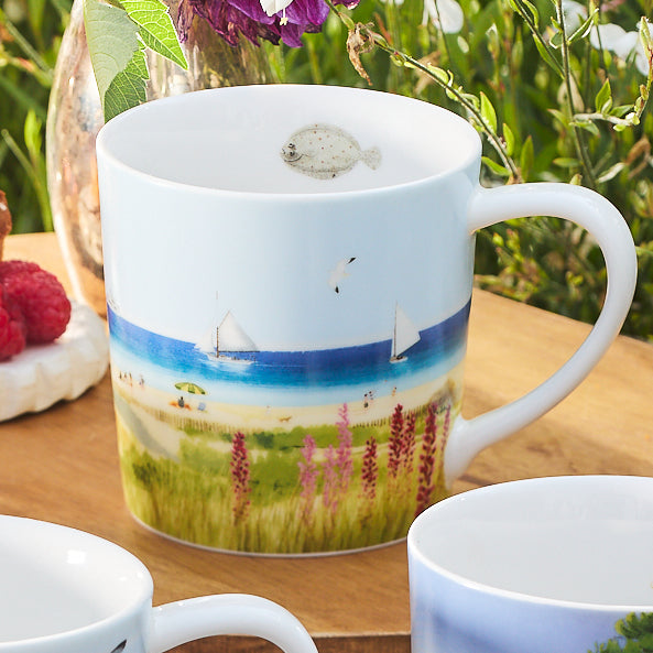 One of the Caskata X Felix Doolittle By the Sea Mugs, from a set of four, is placed on a table next to raspberries and another mug. This white porcelain piece features a beach scene with sailboats, beachgoers, and seagulls. The artwork evokes the gentle charm characteristic of Felix Doolittle's watercolor landscapes.