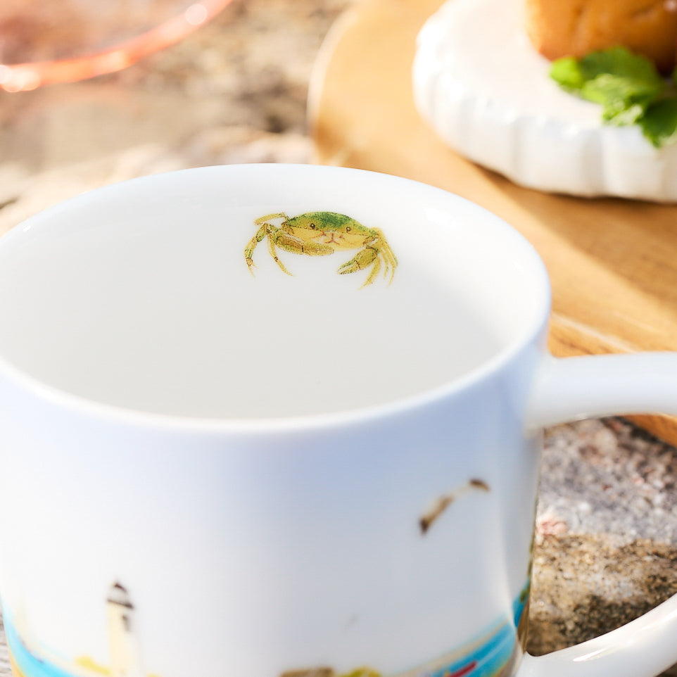 A close-up of a white porcelain mug from the Caskata X Felix Doolittle By the Sea Mugs set, featuring a small green crab illustration on the inside rim in Felix Doolittle&#39;s charming style, sits beside a wooden board with food, garnished with a sprig of parsley.