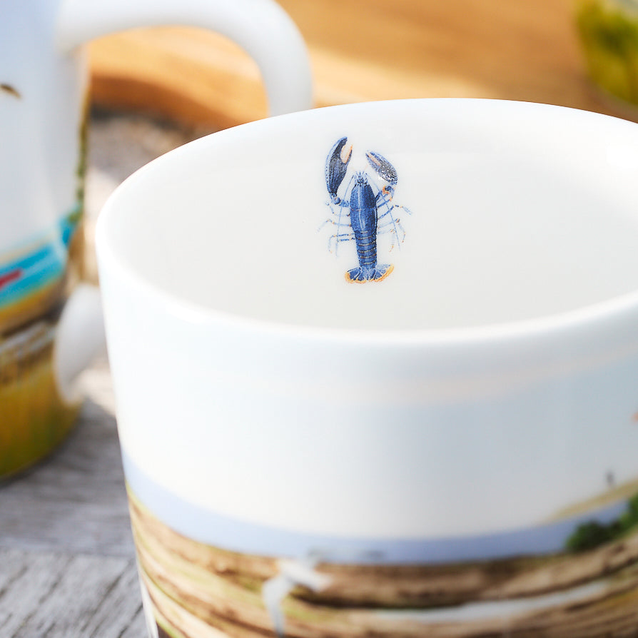 A Caskata X Felix Doolittle By the Sea porcelain mug, adorned with a watercolor landscape on the exterior and an illustration of a blue lobster on the inner bottom surface, showcasing Felix Doolittle's charming style.