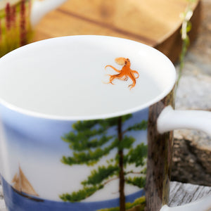 A close-up of a Caskata X Felix Doolittle By the Sea Mug showcases a small illustration of an orange octopus on the inside and an outdoor watercolor landscape with trees and a sailboat on the outside.