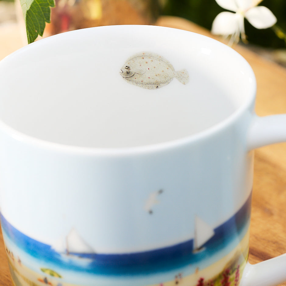 Close-up of a porcelain mug from the Caskata X Felix Doolittle By the Sea Mugs, Set of 4. The mug features a beach scene and a small fish illustration on the inside near the rim. The background is slightly blurred, showing a plant and a white flower. This charming piece evokes the delicate artistry of Felix Doolittle&#39;s watercolor landscapes.