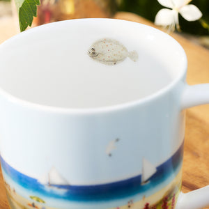 Close-up of a porcelain mug from the Caskata X Felix Doolittle By the Sea Mugs, Set of 4. The mug features a beach scene and a small fish illustration on the inside near the rim. The background is slightly blurred, showing a plant and a white flower. This charming piece evokes the delicate artistry of Felix Doolittle's watercolor landscapes.