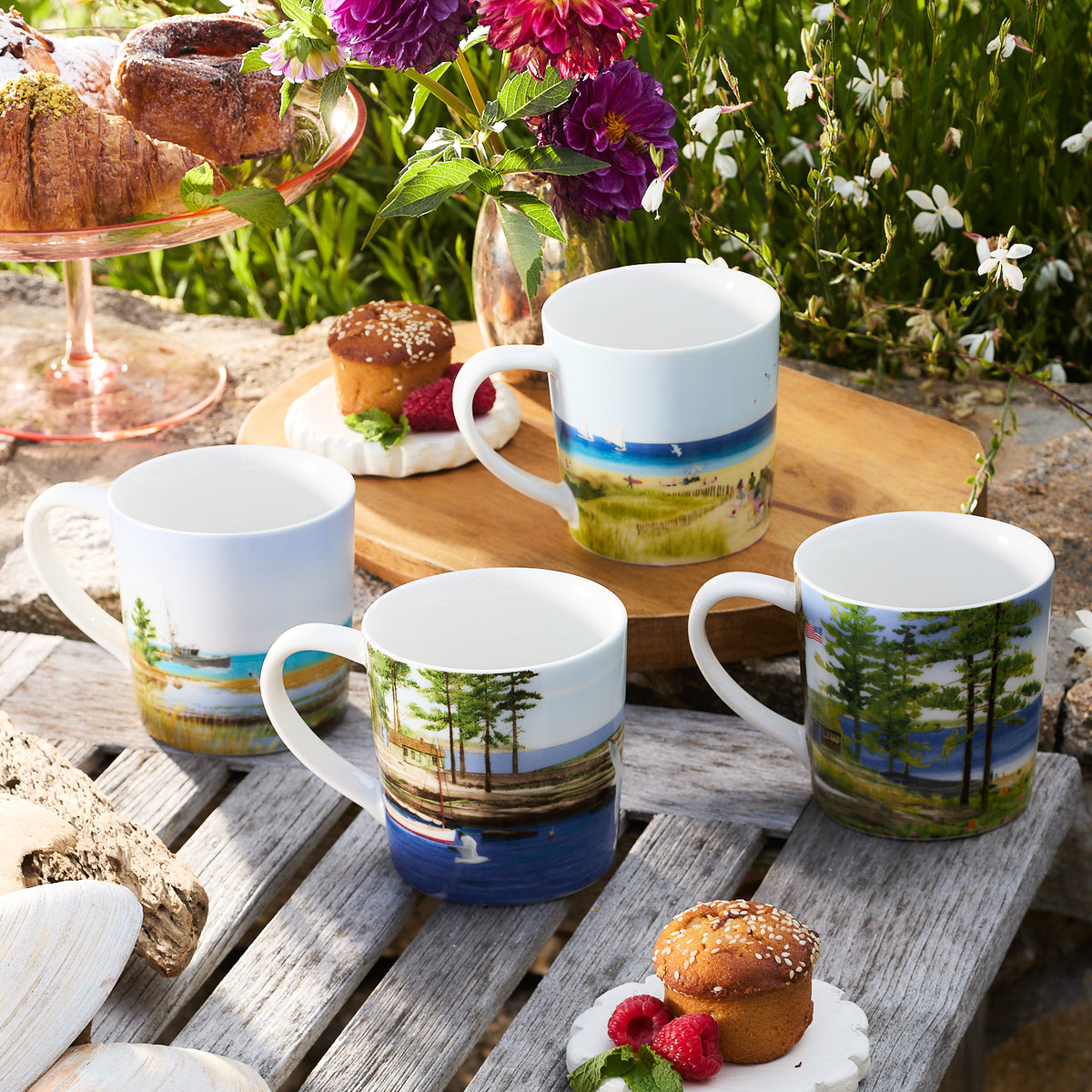 Four Caskata X Felix Doolittle By the Sea Mugs, each adorned with watercolor landscapes by Felix Doolittle, are arranged outdoors on a stone surface with muffins and flowers nearby.