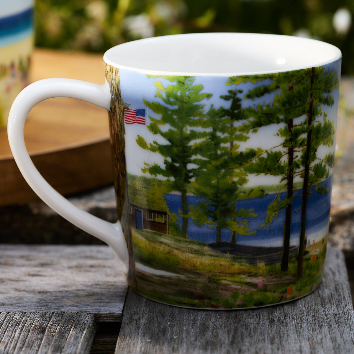 The Caskata X Felix Doolittle By the Sea Mugs, part of Caskata&#39;s charming gift sets, feature a painted scenic landscape with trees, a cabin, a lake, and an American flag, all displayed on rustic wooden surfaces.