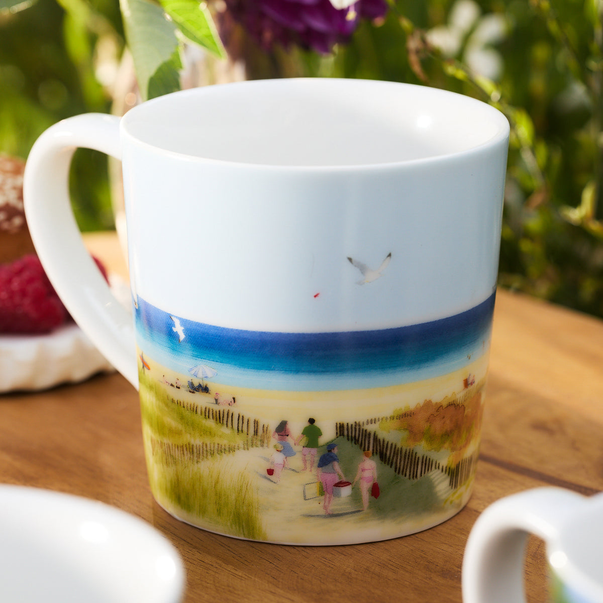 The Caskata X Felix Doolittle By the Sea Mugs, Set of 4 features a white ceramic mug with a watercolor landscape of people walking on a pathway to the beach, surrounded by green fields and a blue ocean in the background. Ideal for gift sets or anyone who appreciates artfully designed porcelain mugs from Caskata.