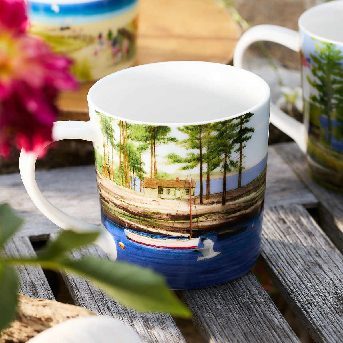 A Caskata X Felix Doolittle By the Sea porcelain mug, part of a set of four, features a scenic watercolor landscape of a lake with a boat, dock, and trees in the background. The mug is placed on a wooden surface next to a flower.