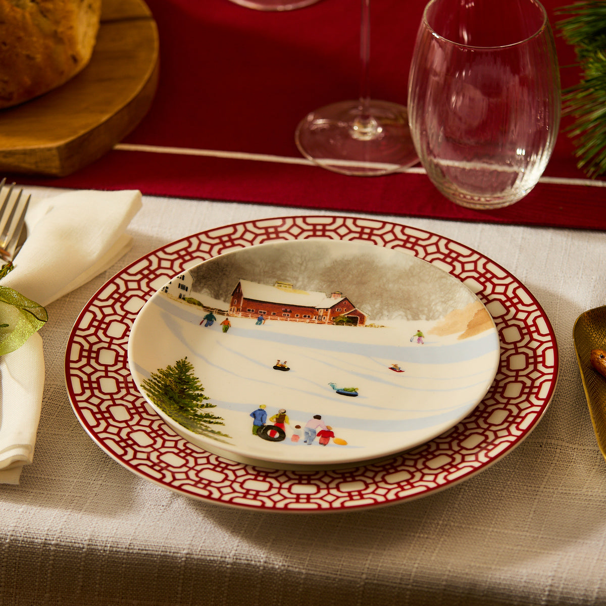 A holiday-themed plate from the Caskata X Felix Doolittle Winter Wonderland set, featuring a watercolor landscape of people sledding, is beautifully placed on a red and white patterned dinner plate on a table set with utensils, napkin, glassware, and bread in the background.
