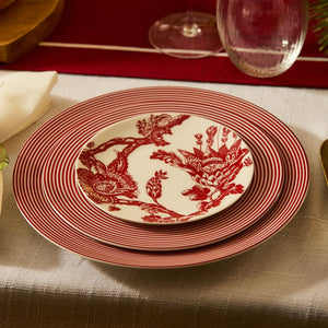 A table setting featuring red and white striped dinner plates with an Arcadia Crimson Small Plate by Caskata Artisanal Home on top, showcasing a beautiful red floral pattern, all crafted from premium porcelain dinnerware. A glass and utensils are visible in the background.