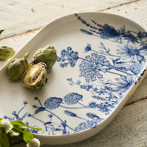 Placed on a rustic wooden surface with scattered leafy greens, the Caskata Summer Blues Large Coupe Oval Platter, crafted from white high-fired porcelain and adorned with a blue floral dinnerware design, holds two whole artichokes and one half artichoke—perfectly capturing the essence of Summer Blues.