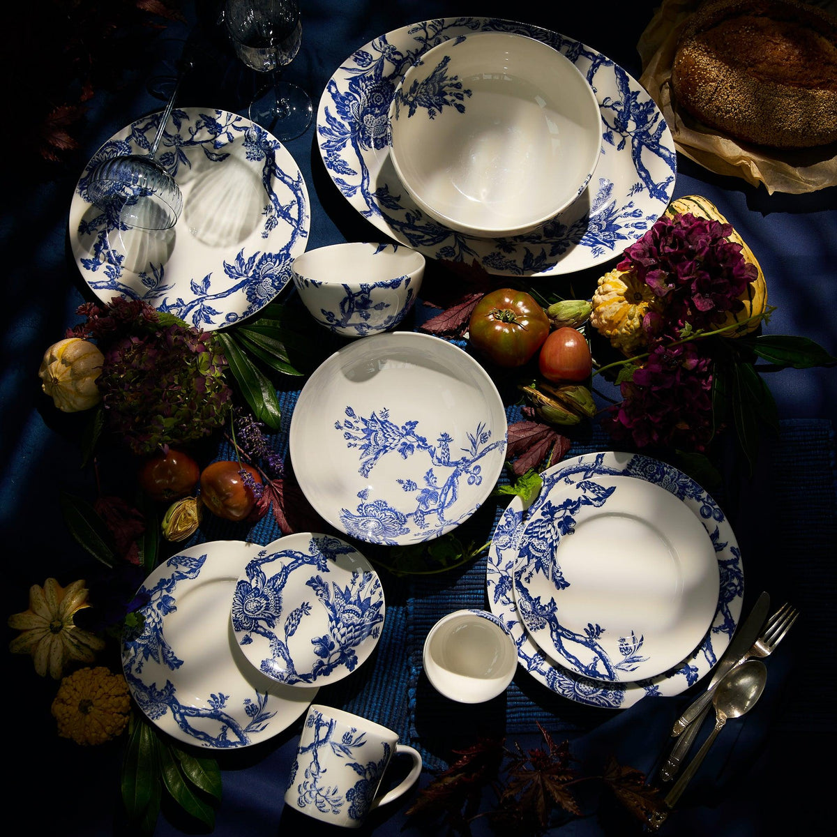 An elegant table setting features the Arcadia Rimmed Dinner Plates by Caskata Artisanal Home, showcasing timeless blue and white graphic florals. The plates are beautifully surrounded by vibrant flowers, ripe fruits, and a freshly baked loaf of bread.