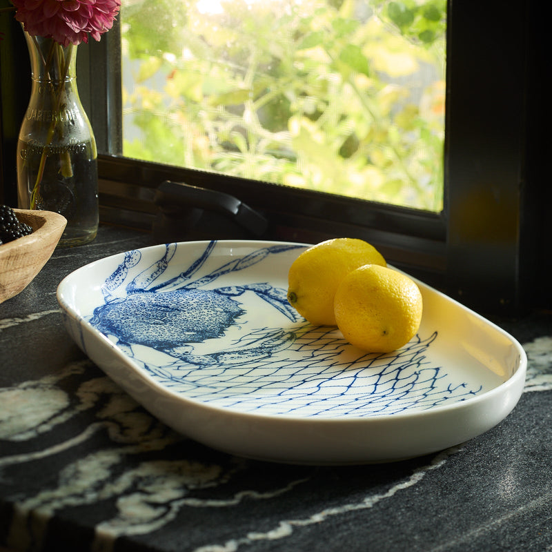 This captivating essential, the Crab Large Coupe Oval Platter by Caskata, boasts a ceramic design featuring a delightful Crabs pattern with two lemons. Made from high-fired porcelain, it is artfully displayed on a marble countertop near a window, alongside a vase and bowl.