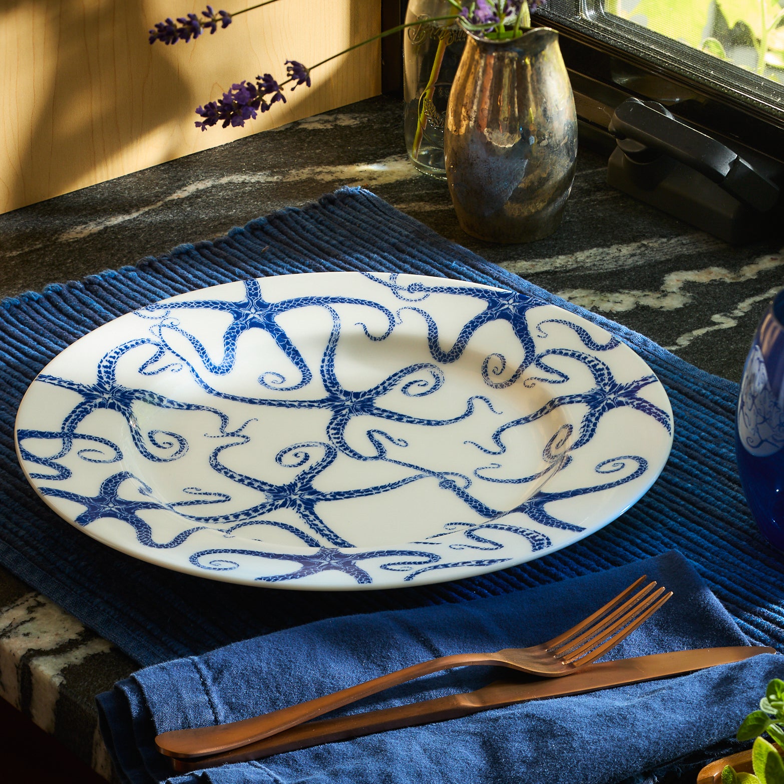 A limited-edition white Starfish Rimmed Dinner Plate from Caskata Artisanal Home features a decorative pattern of blue octopuses intertwined with their tentacles, creating a captivating design.